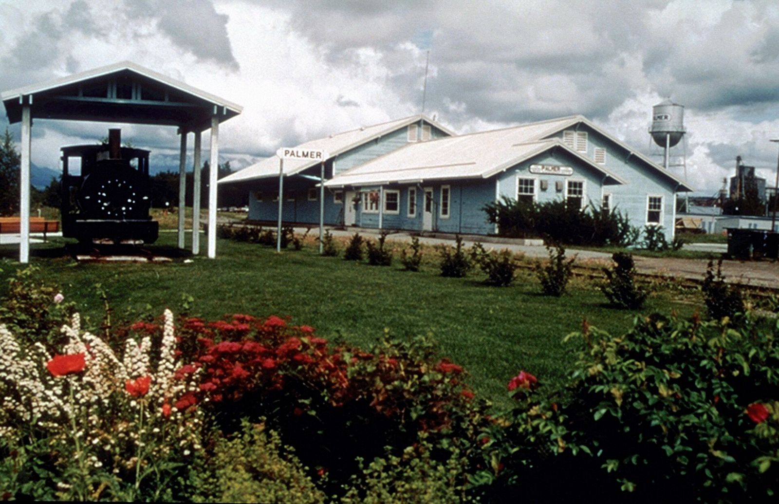 The Palmer train depot, a long white building, beside a historic locomotive and a bed of red flowers