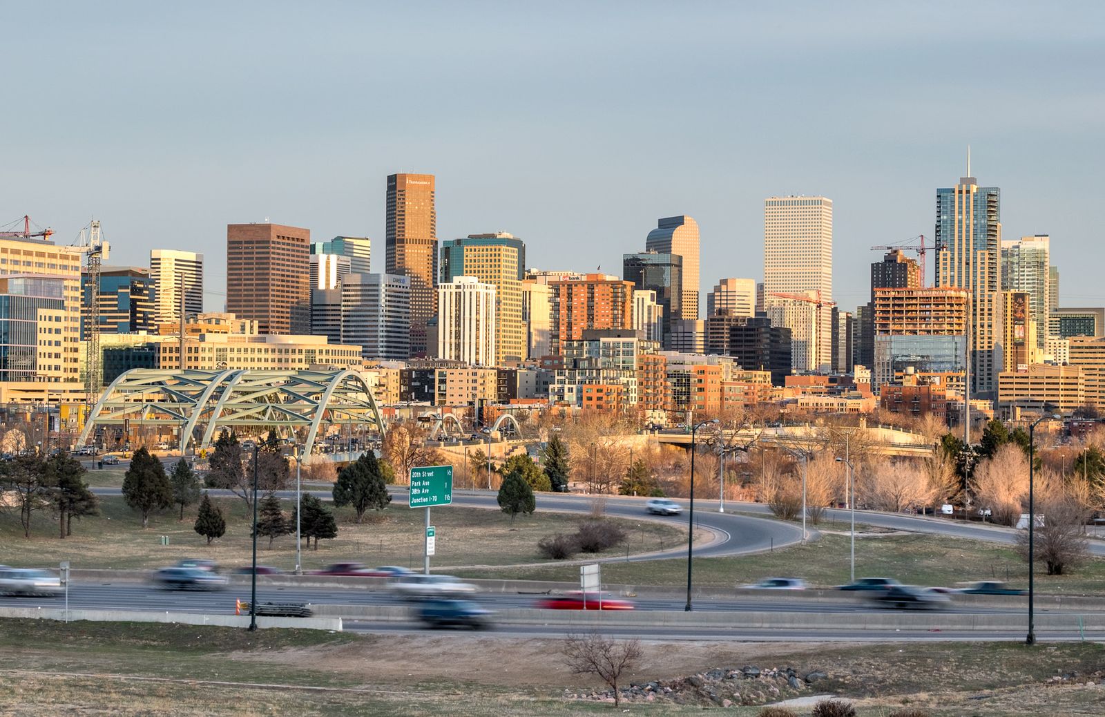 The Denver skyline in warm evening light behind arched pedestrian bridges and lanes of traffic