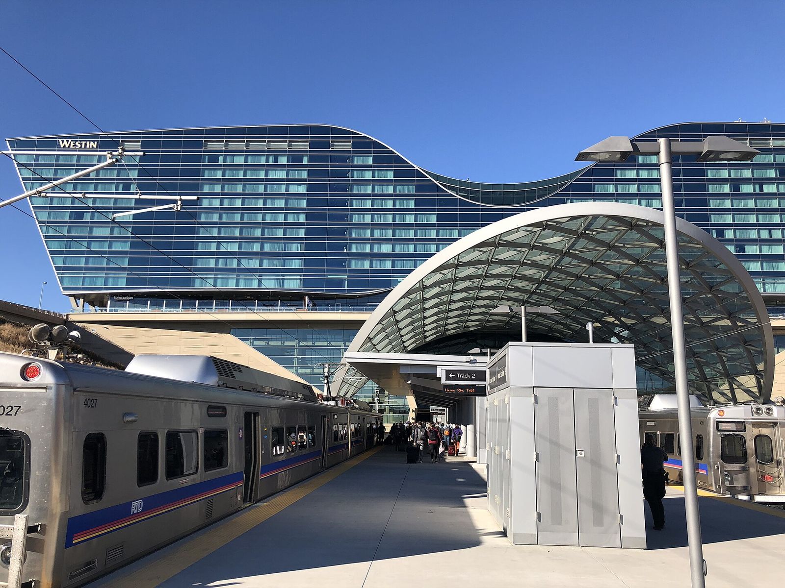 An RTD commuter train at the Denver Airport Station platform, with the curved glass hotel front behind it