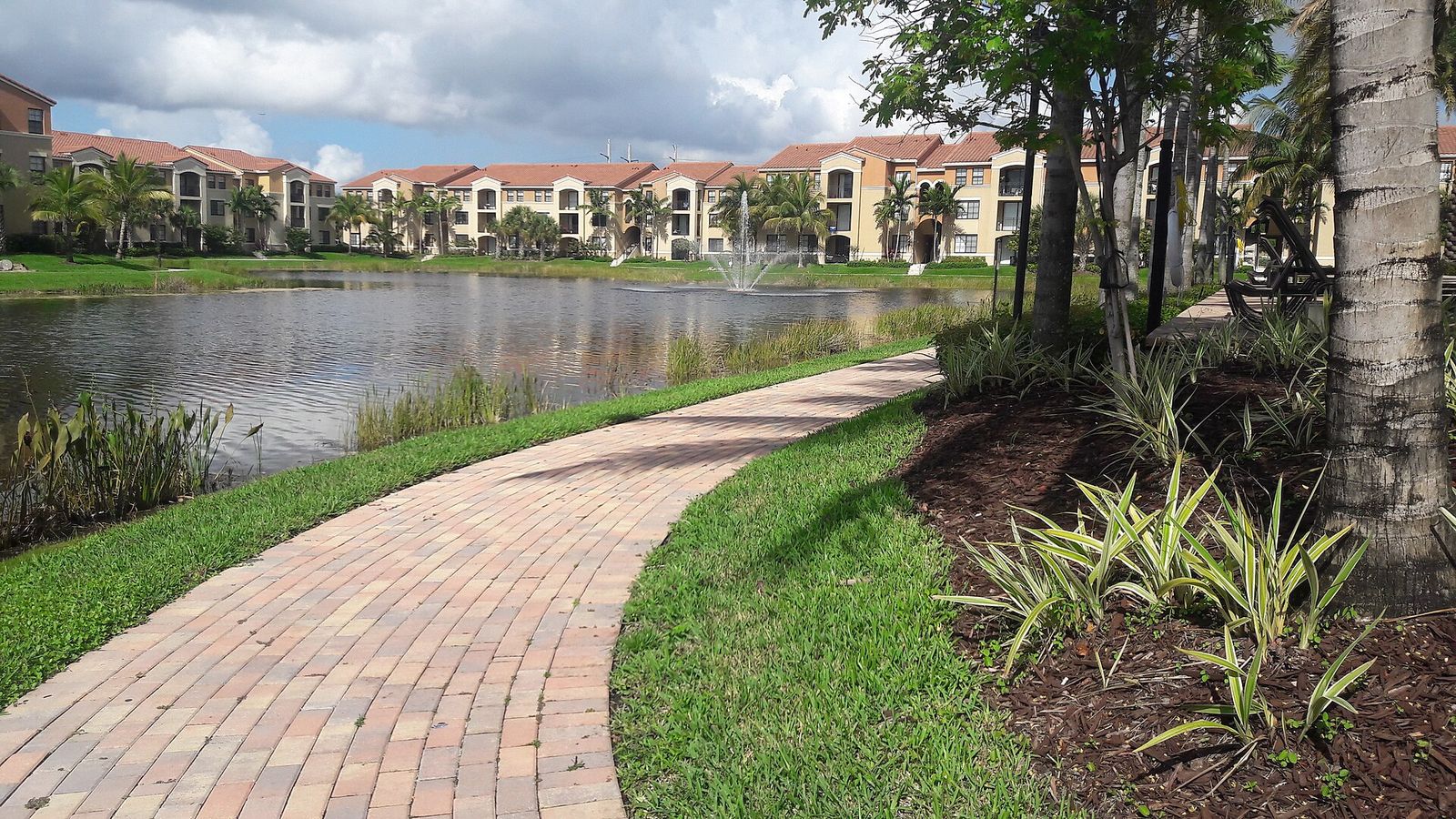 A brick path curving beside a lake and fountain in Doral, with three-story apartment buildings across the water