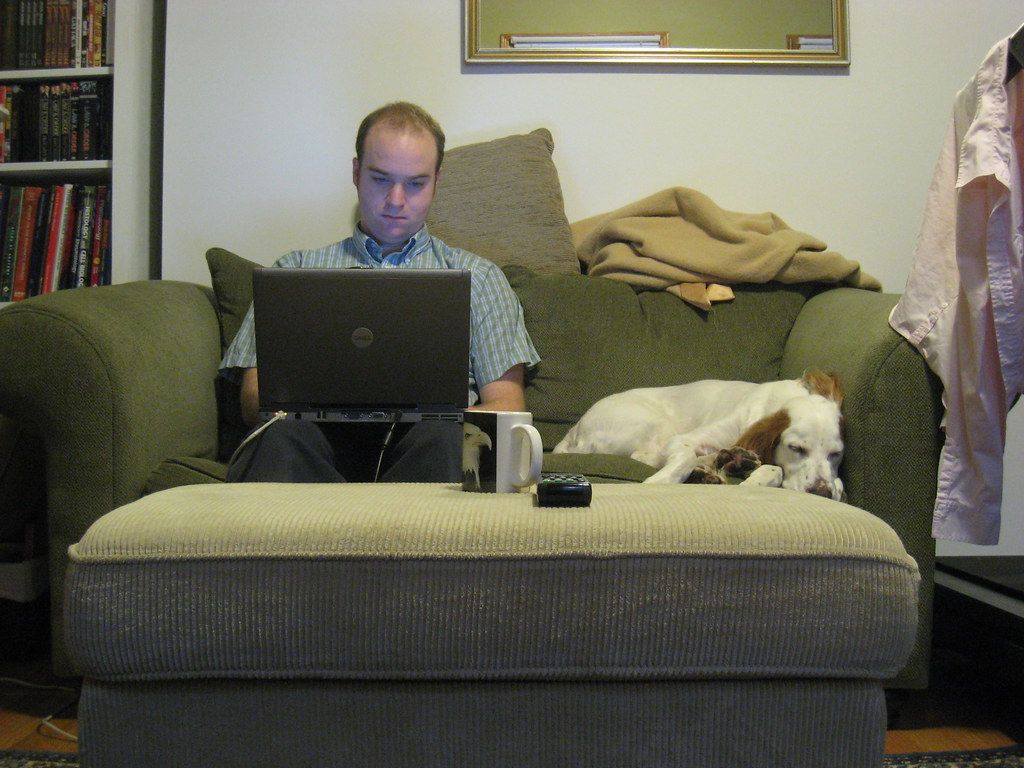 A man working on a laptop on the sofa while his dog sleeps beside him