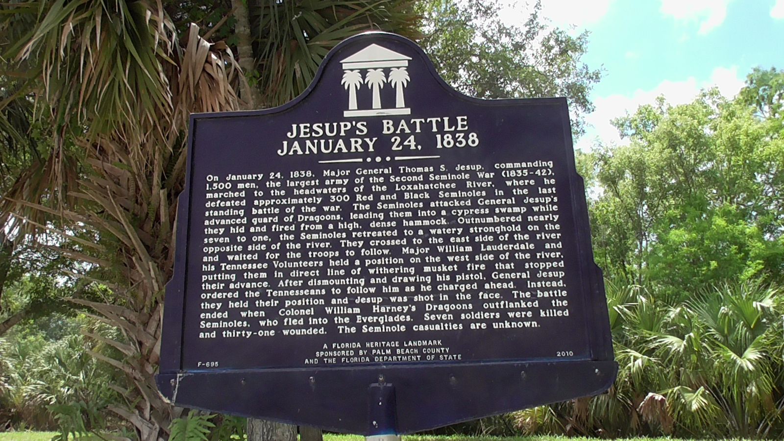A dark Florida historical marker titled Jesup's Battle, January 24, 1838, among palms and shrubs in Jupiter