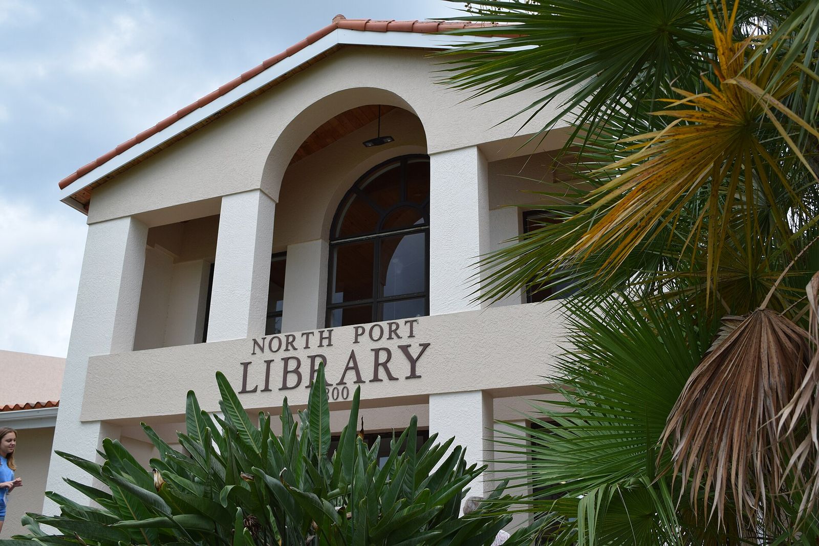 The two-story front of the North Port Public Library, with an arched entry and palms beside it