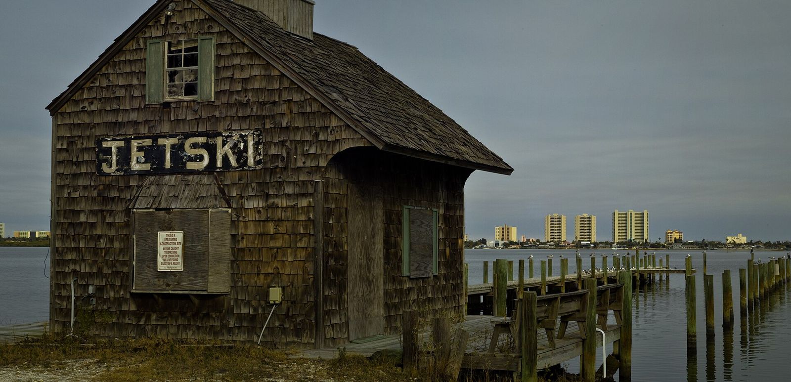 A weathered shingled jet ski shed on the Port Orange riverfront, with condo towers across the water under gray clouds