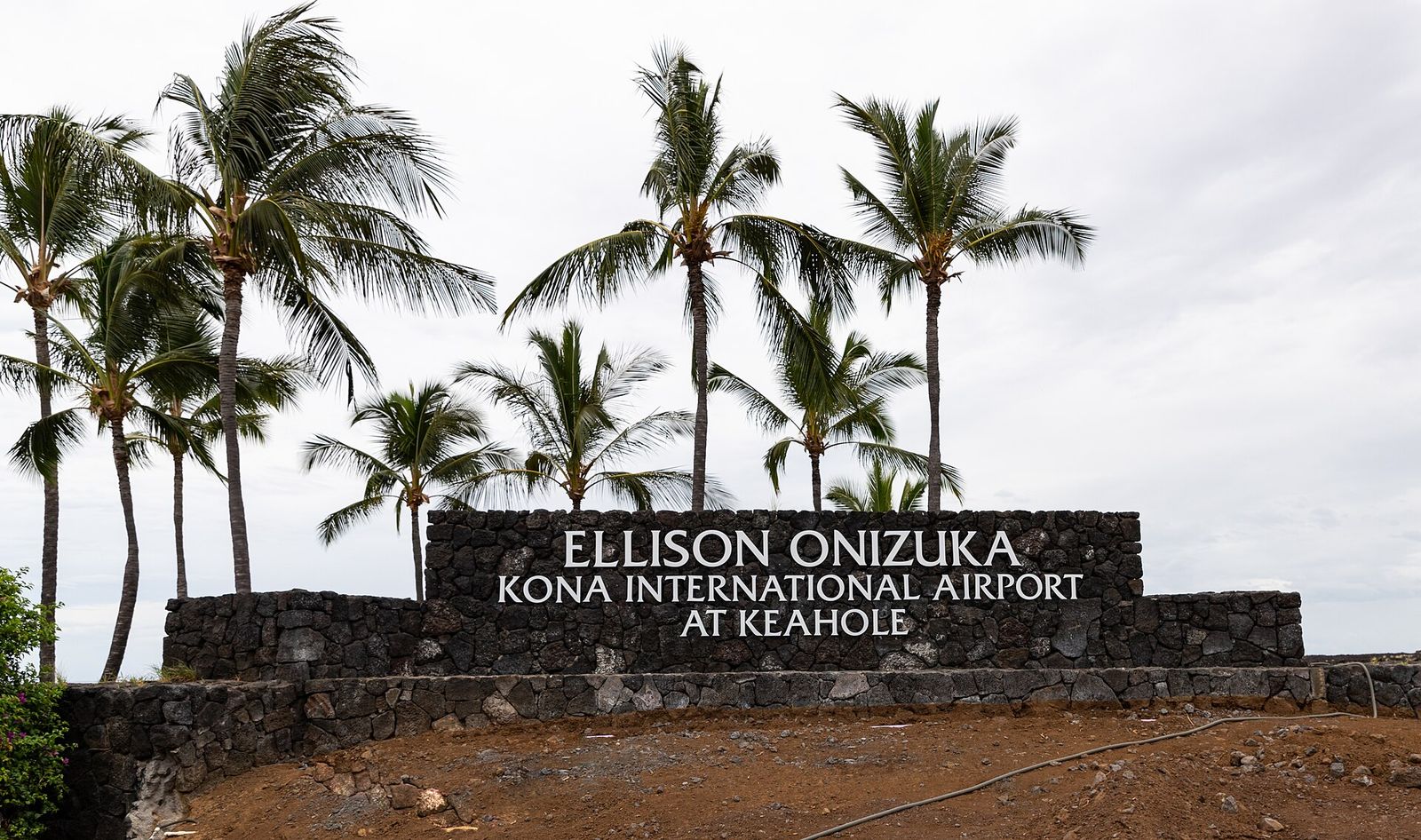 The Ellison Onizuka Kona International Airport at Keahole sign set in a lava rock wall under palm trees