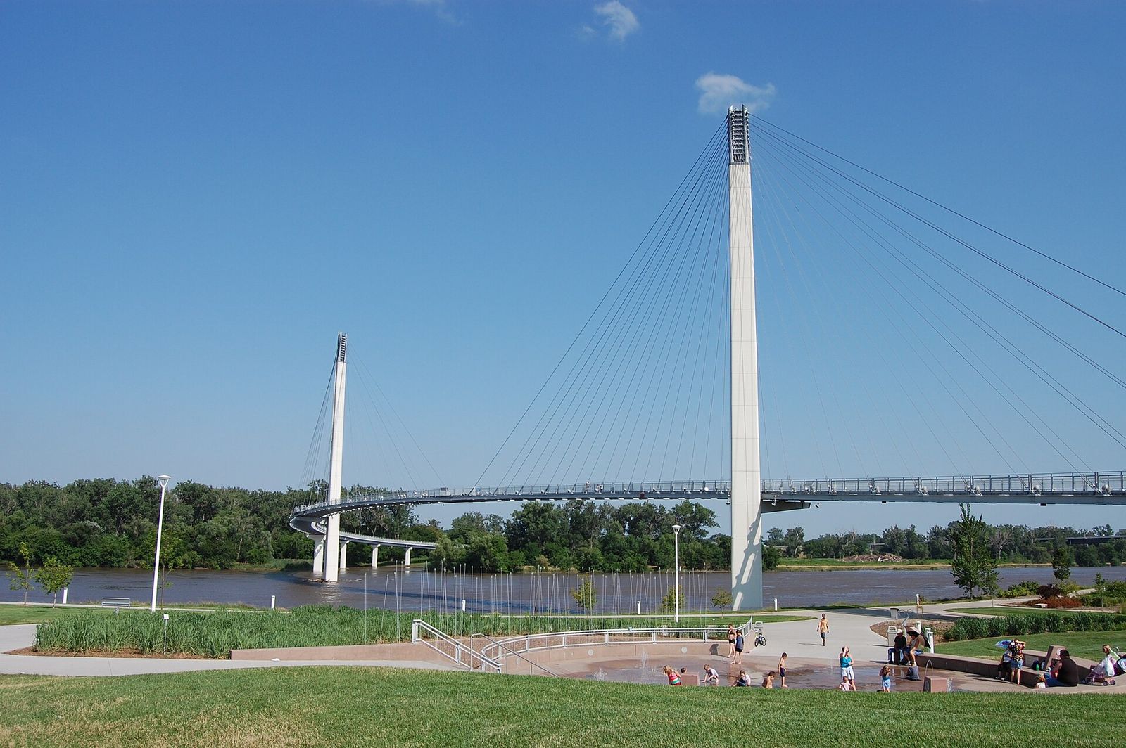 The Bob Kerrey Pedestrian Bridge, with two white towers and fanned cables, crossing the Missouri River beside a riverfront park