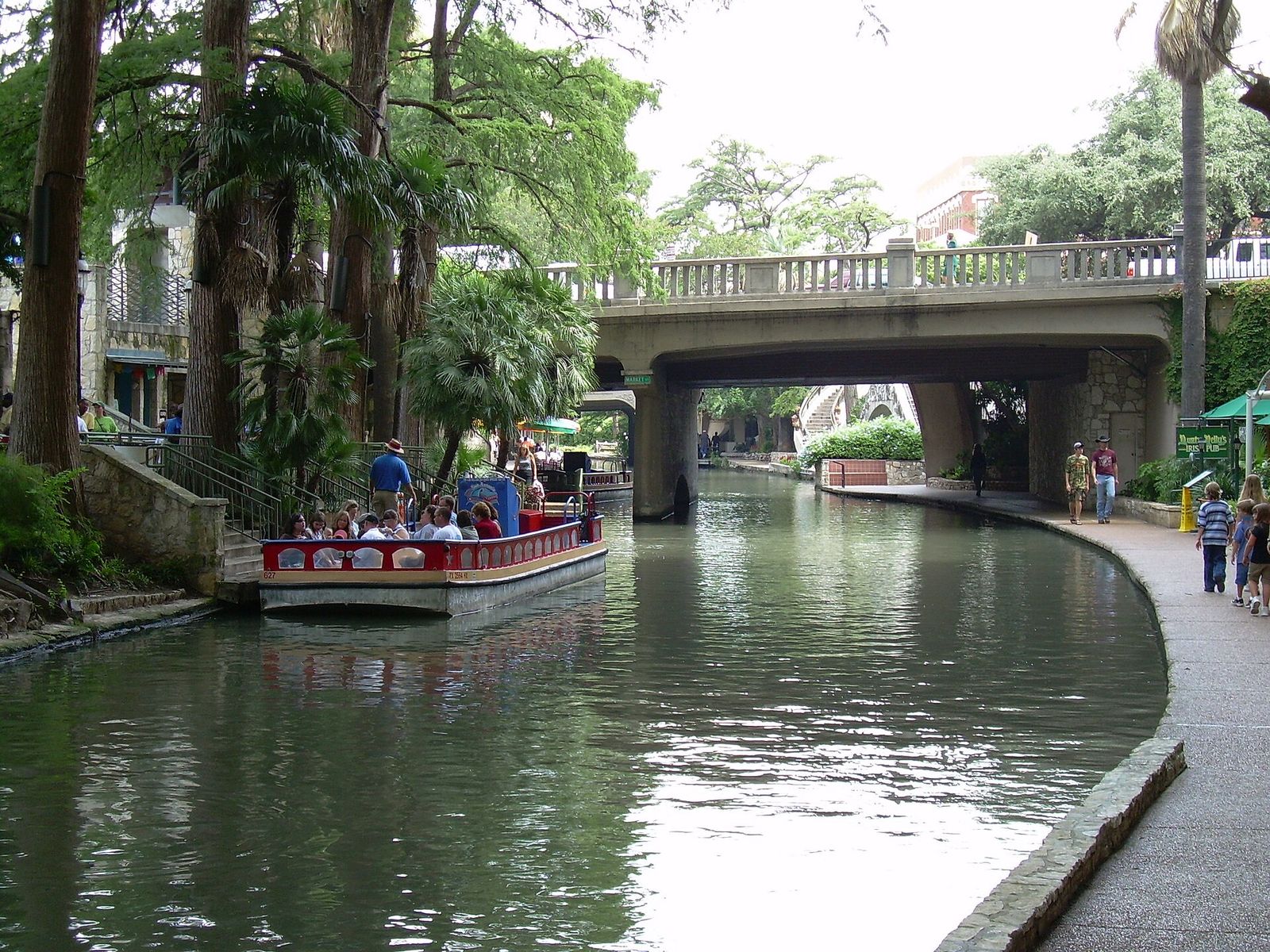 A tour boat carrying passengers along the San Antonio River Walk, with trees and a stone bridge behind it