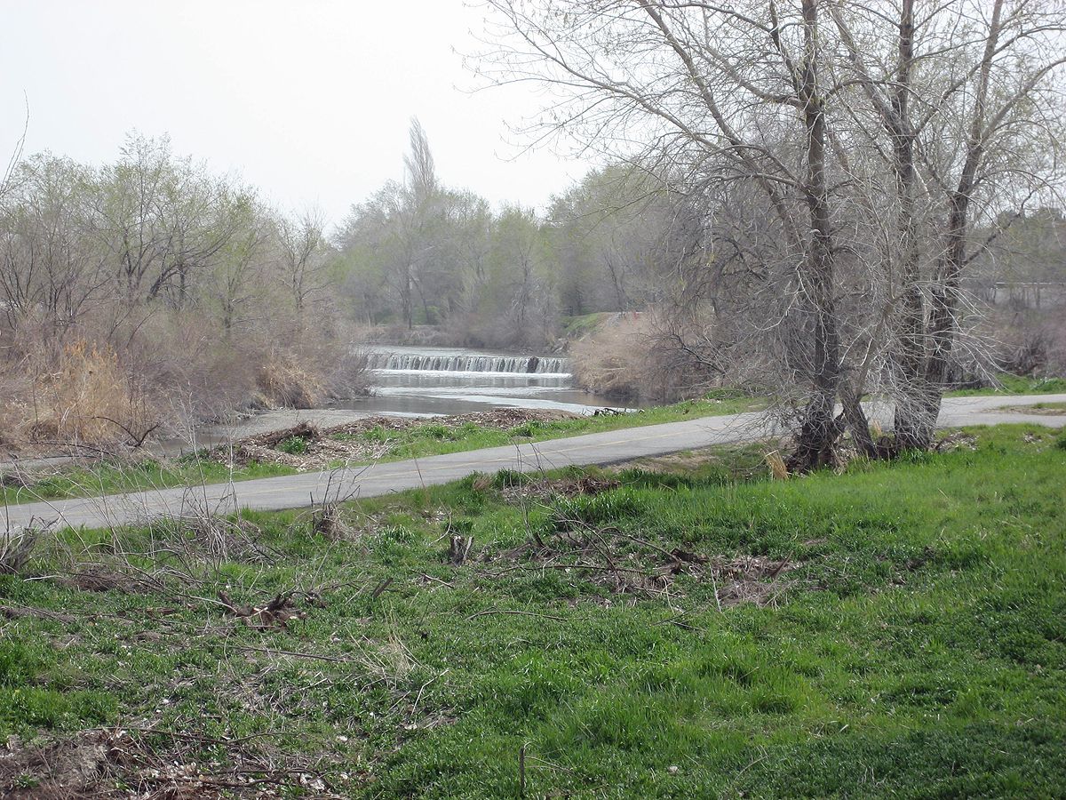 A paved path through spring grass and bare trees beside a small weir on the Jordan River Parkway in Murray