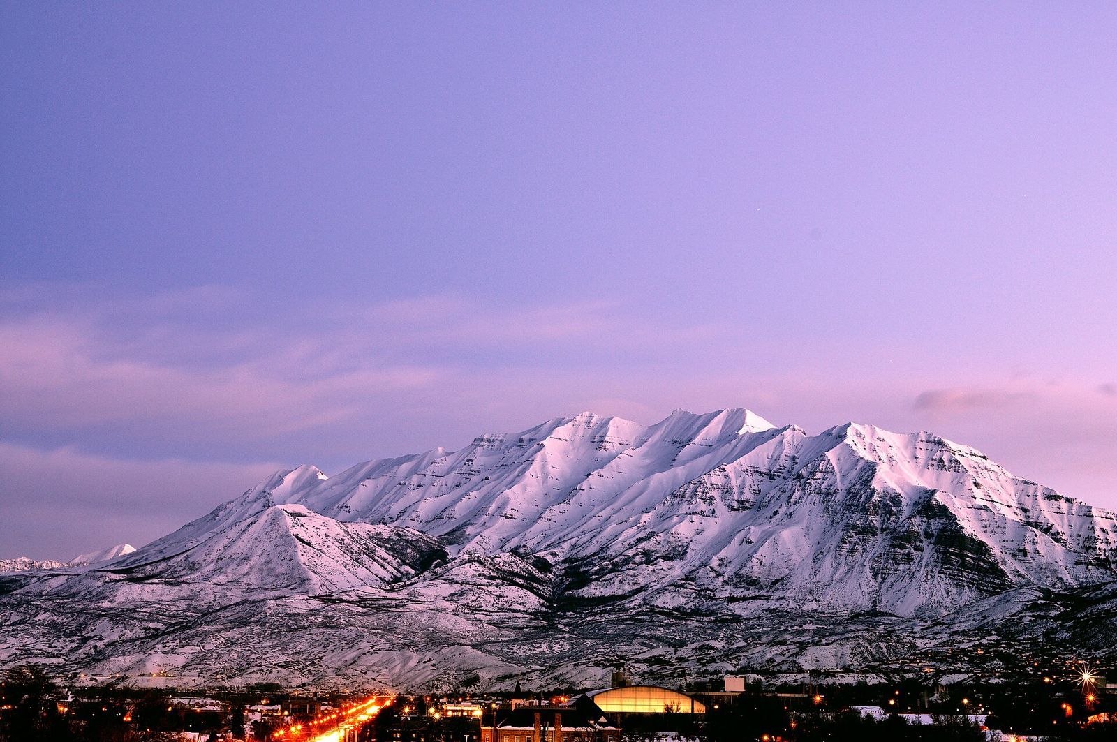 Snow-covered Mount Timpanogos under a pink dawn sky above the lights of Utah Valley