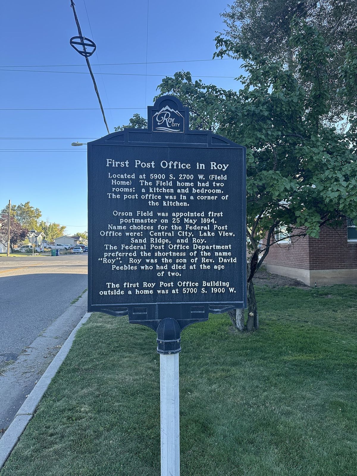 A historical marker describing the first post office in Roy, beside a lawn and a street